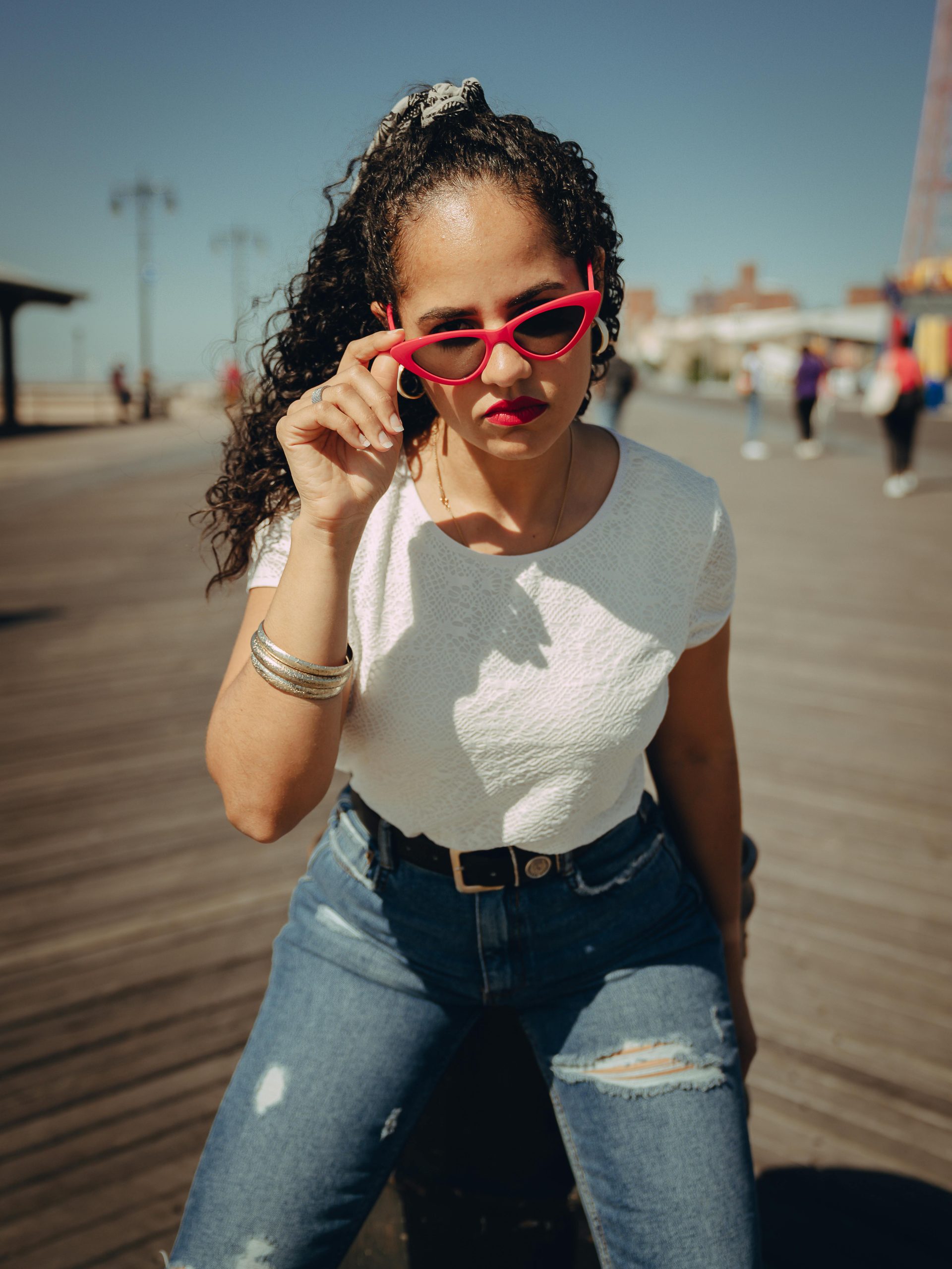 Stylish woman with red sunglasses poses confidently on a sunny NY boardwalk.